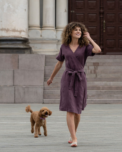 A woman with medium-length curly hair smiles and poses, wearing a shadow purple, short-sleeved wrap dress with a tie at the waist. 