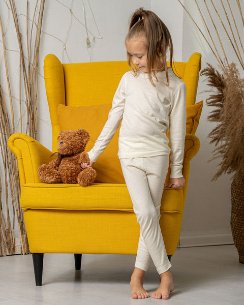 A girl standing by a yellow armchair, holding a teddy bear by the hand, wearing the menique Kids' 160 Merino Pants in Natural color.
