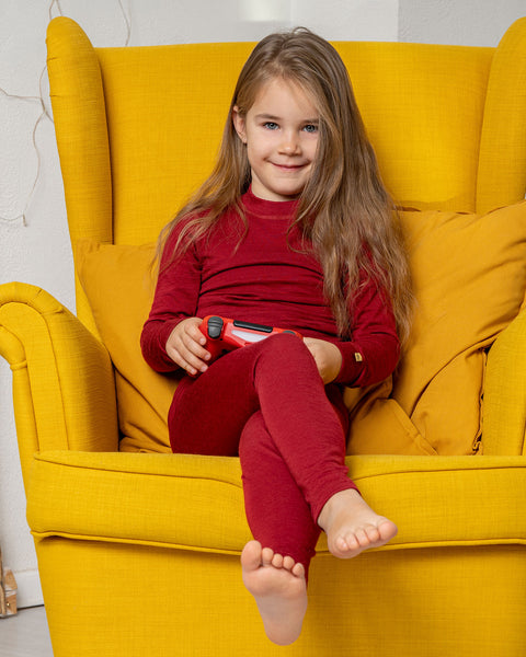 A girl, sitting in a yellow armchair, holding a game controller wearing the menique Kids' 160 Merino Pants in Royal Cherry color. 