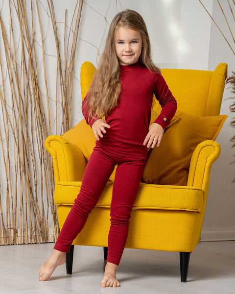 A girl, standing by a yellow armchair, wearing the menique Kids' 160 Merino Pants in Royal Cherry color, with one leg slightly to the side and both hands on hips. 