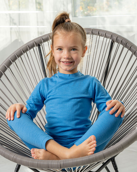 A girl sitting in a longe chair, with her legs crossed and her hands on her knees. She is wearing the merino 160gsm long sleeve top in light blue color and matching pants. 
