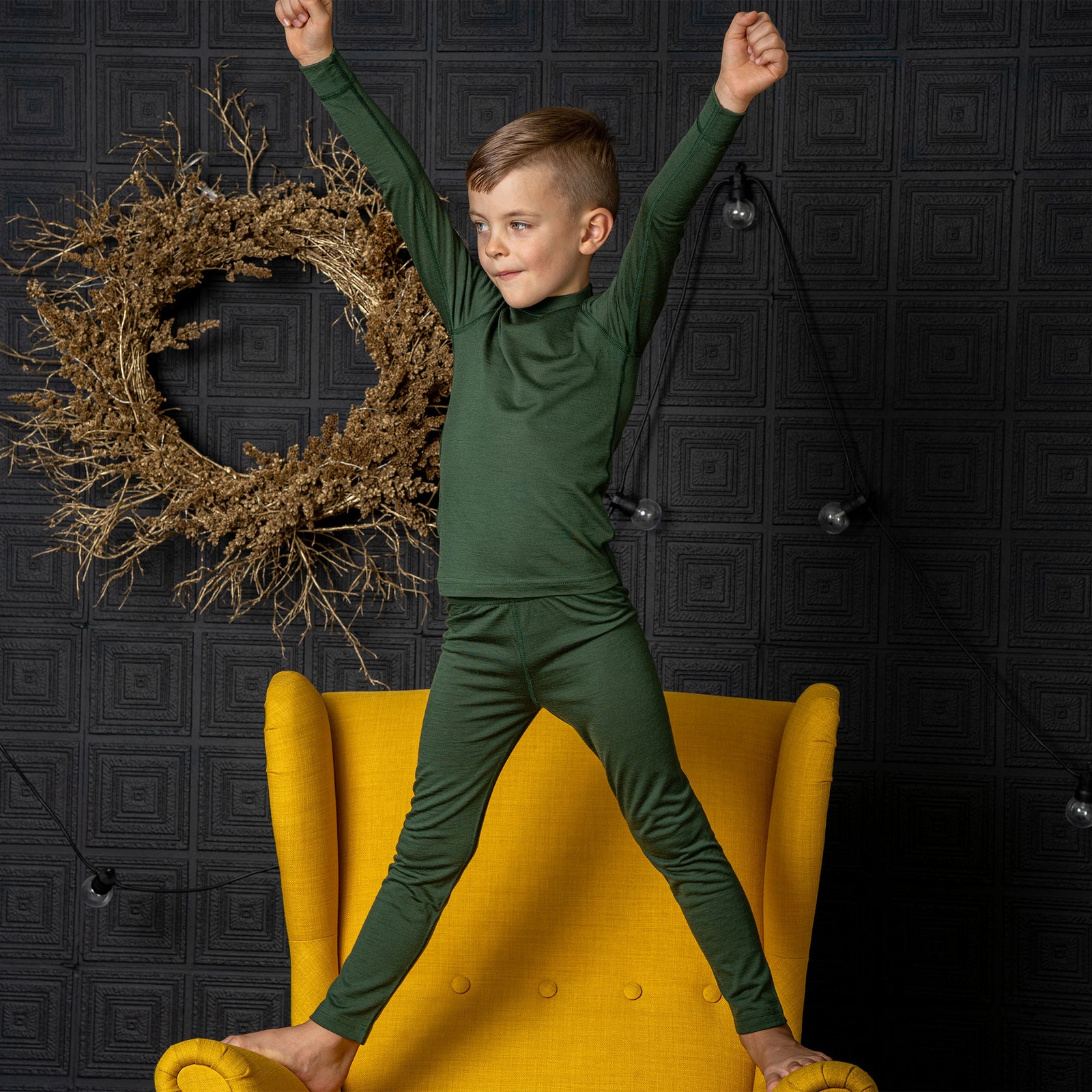Een jongen zit op een stoel, met één been erop gelegd, en draagt de Menique Kids' 160 Merino Broek in donkergroene kleur. Hij laat zijn hoofd rusten op de knie van het opgetrokken been.