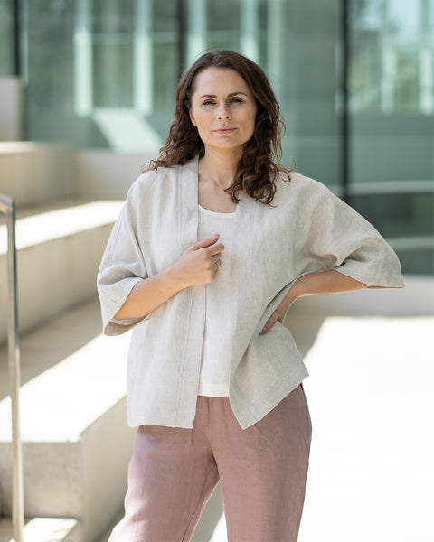 A woman with brown hair, wearing a light beige kimono-style jacket over mauve trousers, stands on indoor concrete stairs.