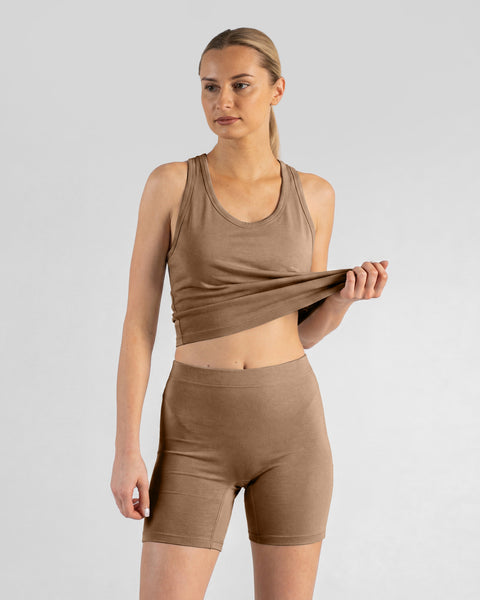 A model in a three-quarter view poses against a light gray background, wearing a matching beige tank top and bike shorts. She is gently holding the hem of her tank top up with her left hand.