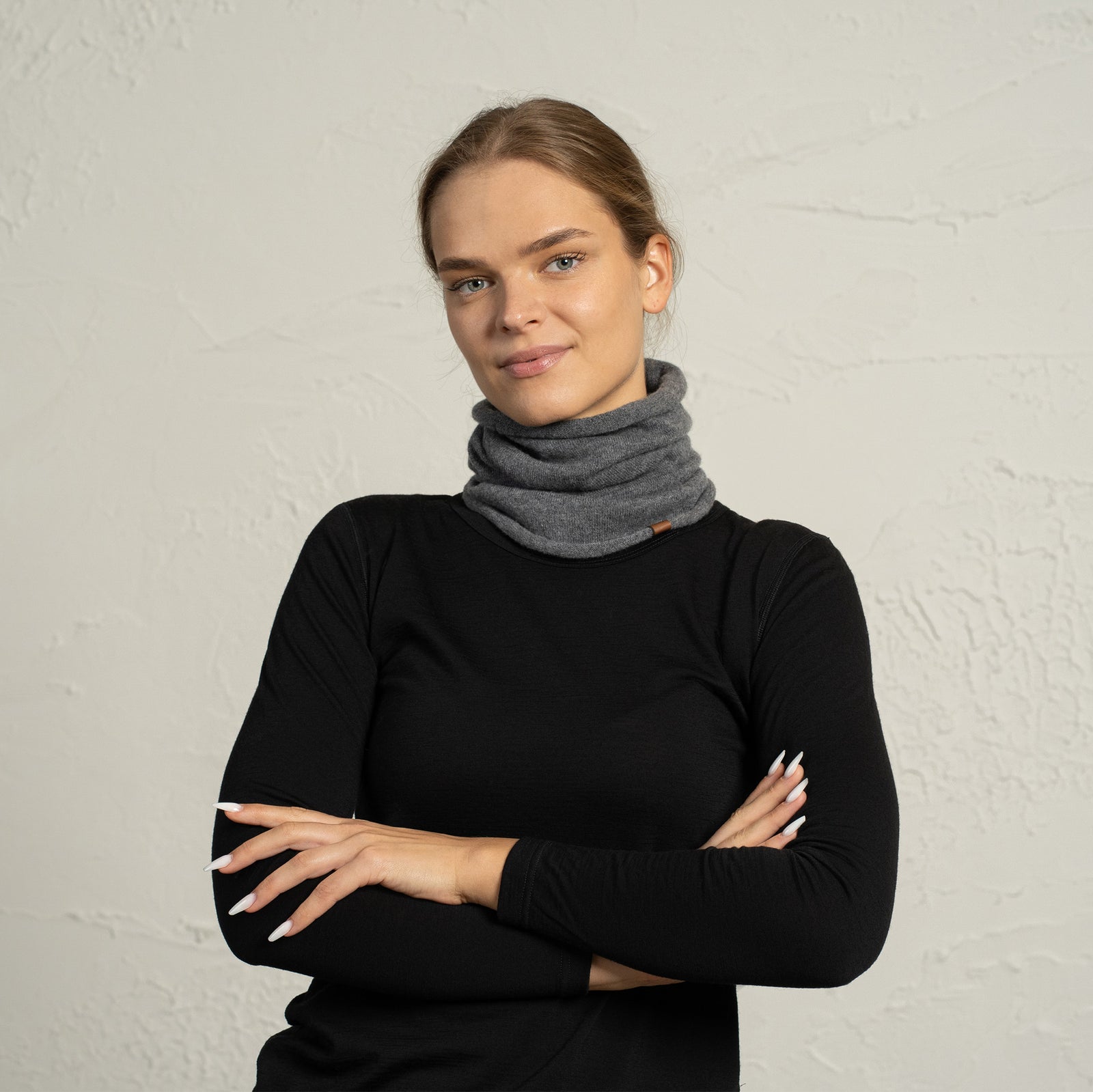 Vrouw draagt een beige merinowollen nekwarmer, staat met gekruiste armen en kijkt naar de camera.