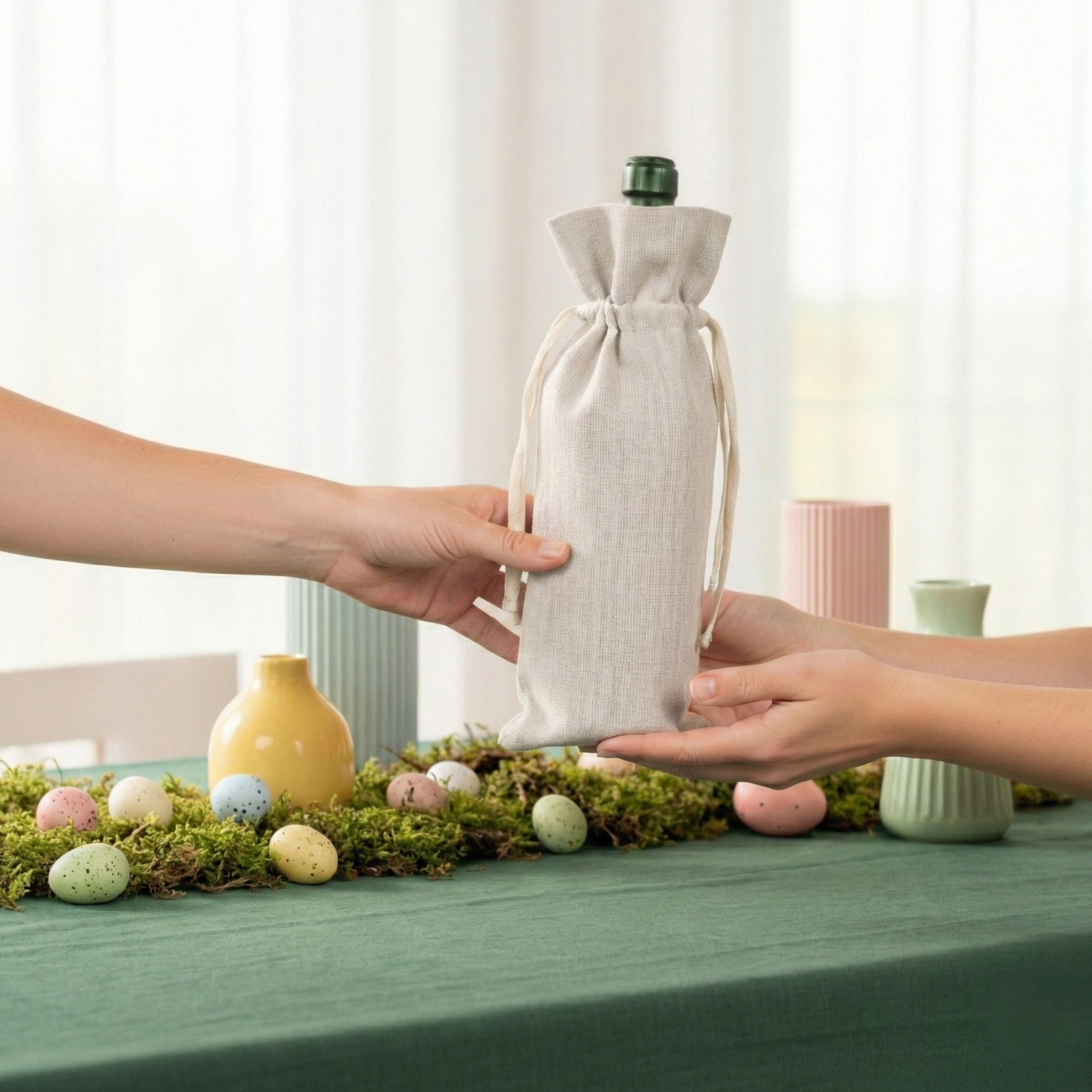 Drie linnen cadeauzakken voor wijnflessen in rood, groen en natuurlijke tinten, tentoongesteld op een lentetafel met bloemen, kaarsen en pastelkleurige versiering.