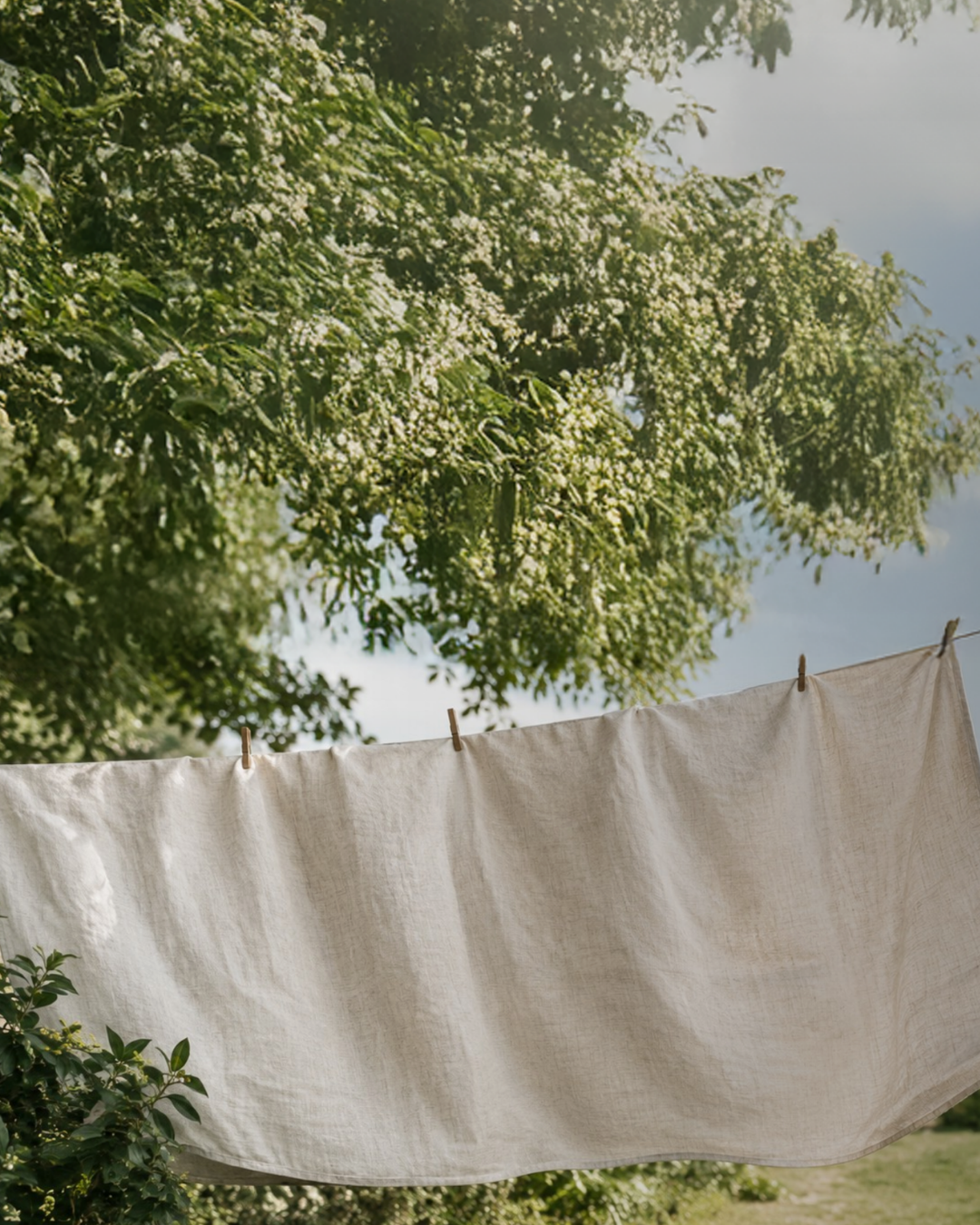 A large, light-colored natural linen cloth hanging on an outdoor clothesline, secured with wooden clothespins and blowing gently in the breeze. The background features a lush green yard with trees and a stone building visible on the right side.