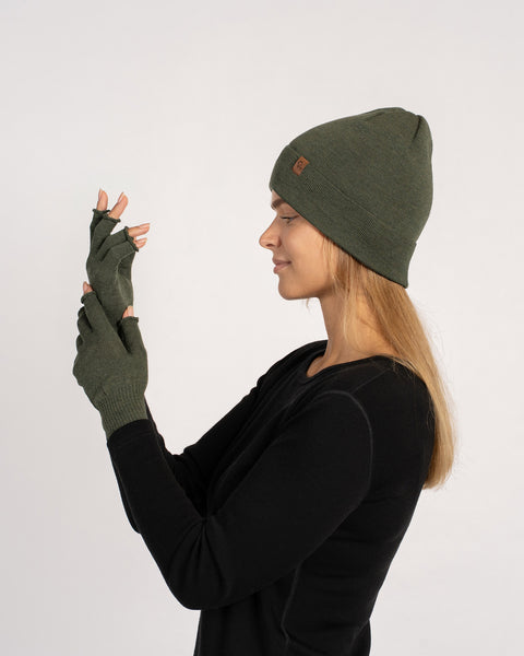 Zijaanzicht van een vrouw die een donkergroene gebreide muts en bijpassende vingervrije handschoenen draagt, terwijl ze haar opgeheven handen bekijkt.