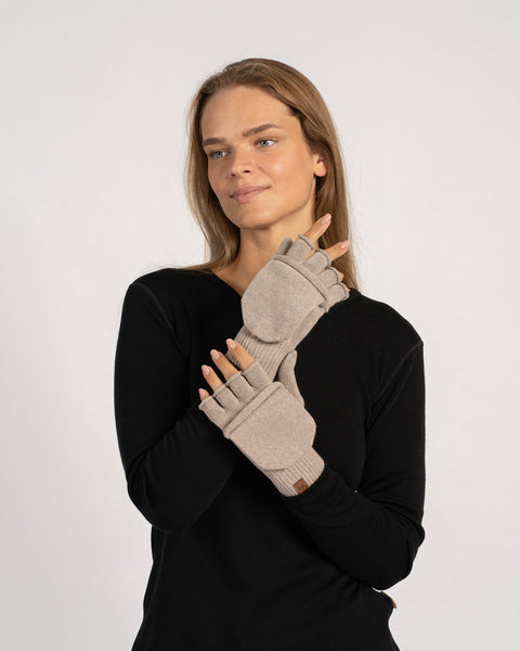 Three-quarter view of the model pulling on a creamy-beige convertible mitten with the flap tucked, arms slightly raised.