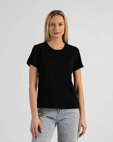 A studio portrait of a blonde woman standing and looking directly at the camera with her arms relaxed by her sides. She is wearing a regular fit, solid black short-sleeved crew-neck t-shirt and light-wash blue jeans. The background is a solid light grey.