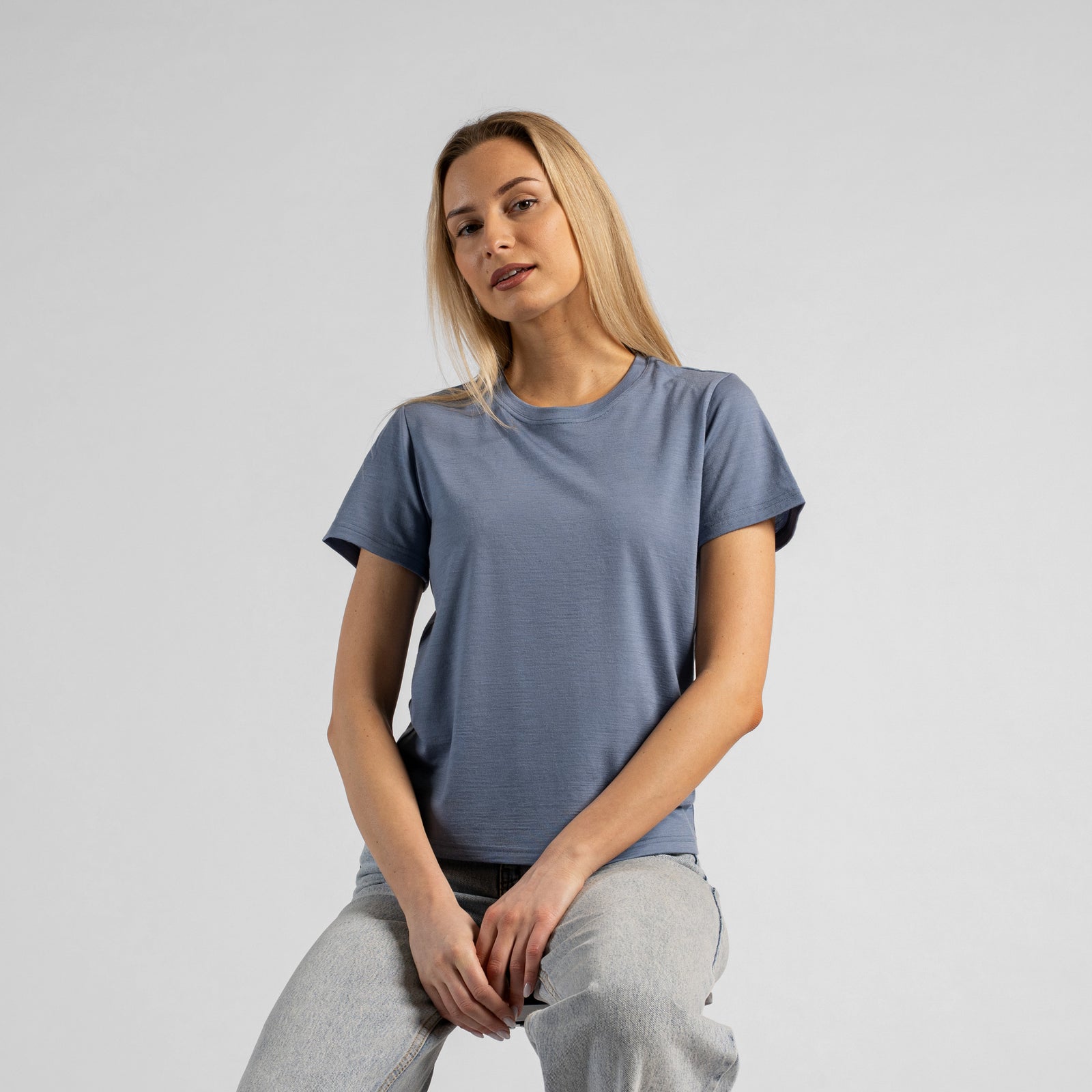 A studio portrait of a blonde woman standing and looking directly at the camera, with her left hand casually tucked into the front pocket of her light-wash blue jeans. She is wearing a regular fit, charcoal grey short-sleeved crew-neck t-shirt against a solid light grey background.