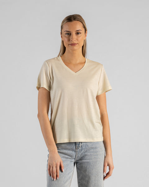A studio portrait of a blonde woman standing and looking directly at the camera with her arms relaxed by her sides. She is wearing a regular fit, light cream natural-colored short-sleeved v-neck t-shirt and light-wash blue jeans against a plain light grey background.