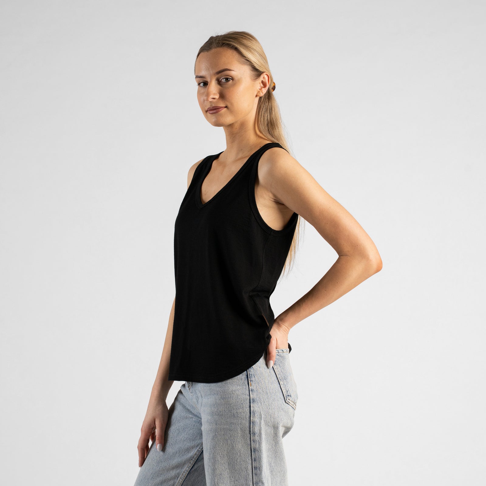 A studio portrait of a blonde woman sitting on a black stool and leaning slightly forward. Her hands are resting on her thighs just above her knees. She is wearing a regular fit, dusty blue sleeveless v-neck top and light-wash blue jeans. The background is a solid light grey.