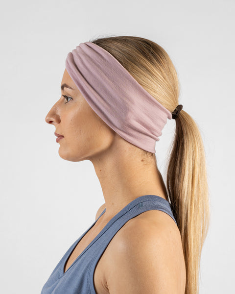 A side profile studio portrait of a blonde woman facing left against a plain light grey background. She is wearing a dusty blue sleeveless top and a dusty pink ribbon headband over her hair, which is tied back in a ponytail.