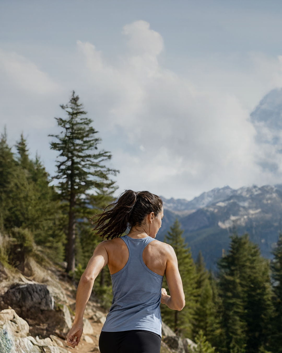 A woman with a dark ponytail running on a mountain trail, wearing a light blue athletic tank top. Pine trees and a mountain range are visible in the background under a cloudy sky.