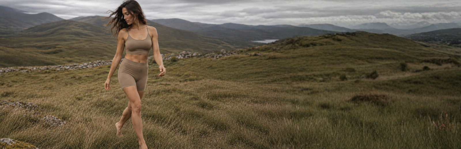 A woman with long wavy hair walks through a grassy landscape wearing a tan sports bra and matching boy shorts. The scene is set against a dramatic backdrop of rolling hills and a dark, heavily clouded sky.