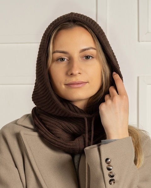 A blonde woman looking directly at the camera, wearing a dark brown ribbed knit balaclava hood tucked into a beige coat.