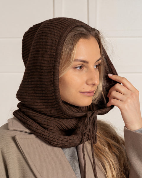 A woman in a three-quarter profile adjusting the hood of a dark brown ribbed knit balaclava featuring matching drawstrings.