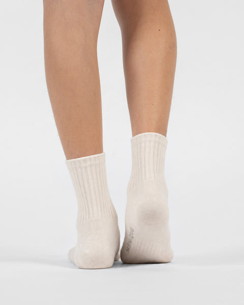 A back view of a person's lower legs and feet wearing natural off-white crew socks, with the right heel slightly lifted to show the "MENIQUE" logo on the sole.