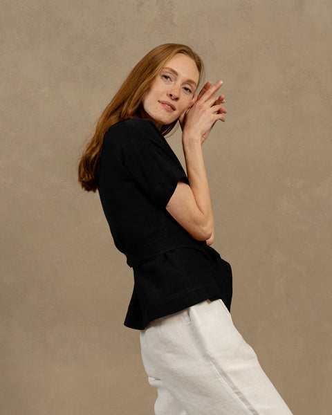 A fair-skinned person with long, auburn hair, wearing menique black V-neck linen wrap top with short, wide sleeves and light-colored pants. They are holding the ends of the top's tie in front of them, forming a bow.