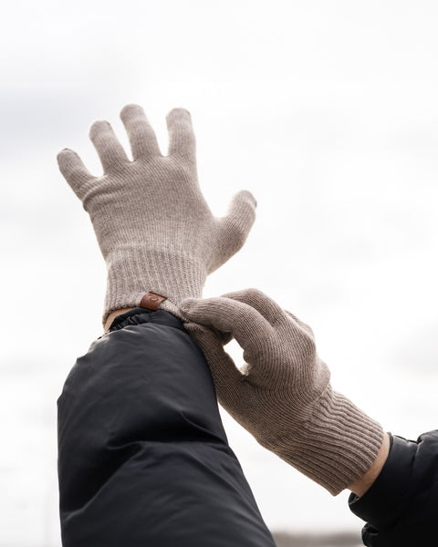 A close-up shot of a person's hands putting on merino wool knit gloves outdoors against a blurred, overcast sky. The gloves are ribbed at the wrists and have a small brown tag. The person is wearing a dark blue or black jacket.