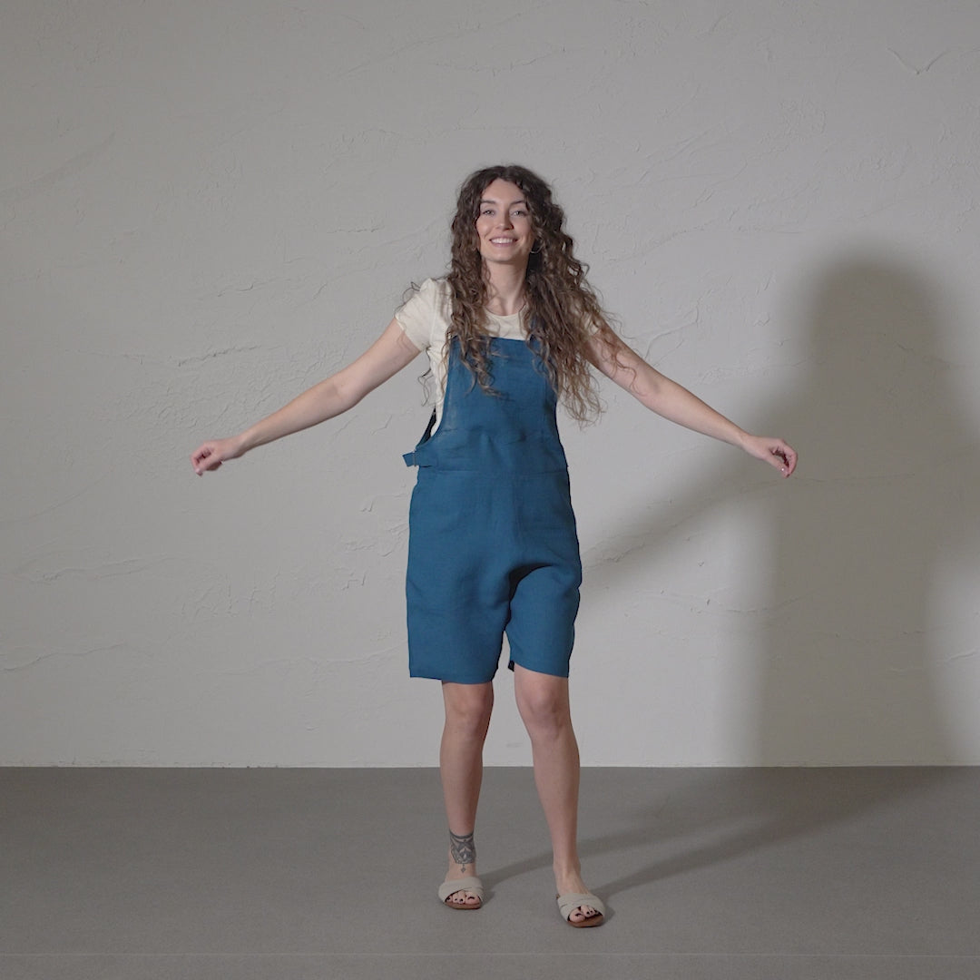 A woman with long, curly brown hair dances energetically in a short video, wearing a white t-shirt underneath short linen romper. She points with both hands and moves against a plain studio backdrop.