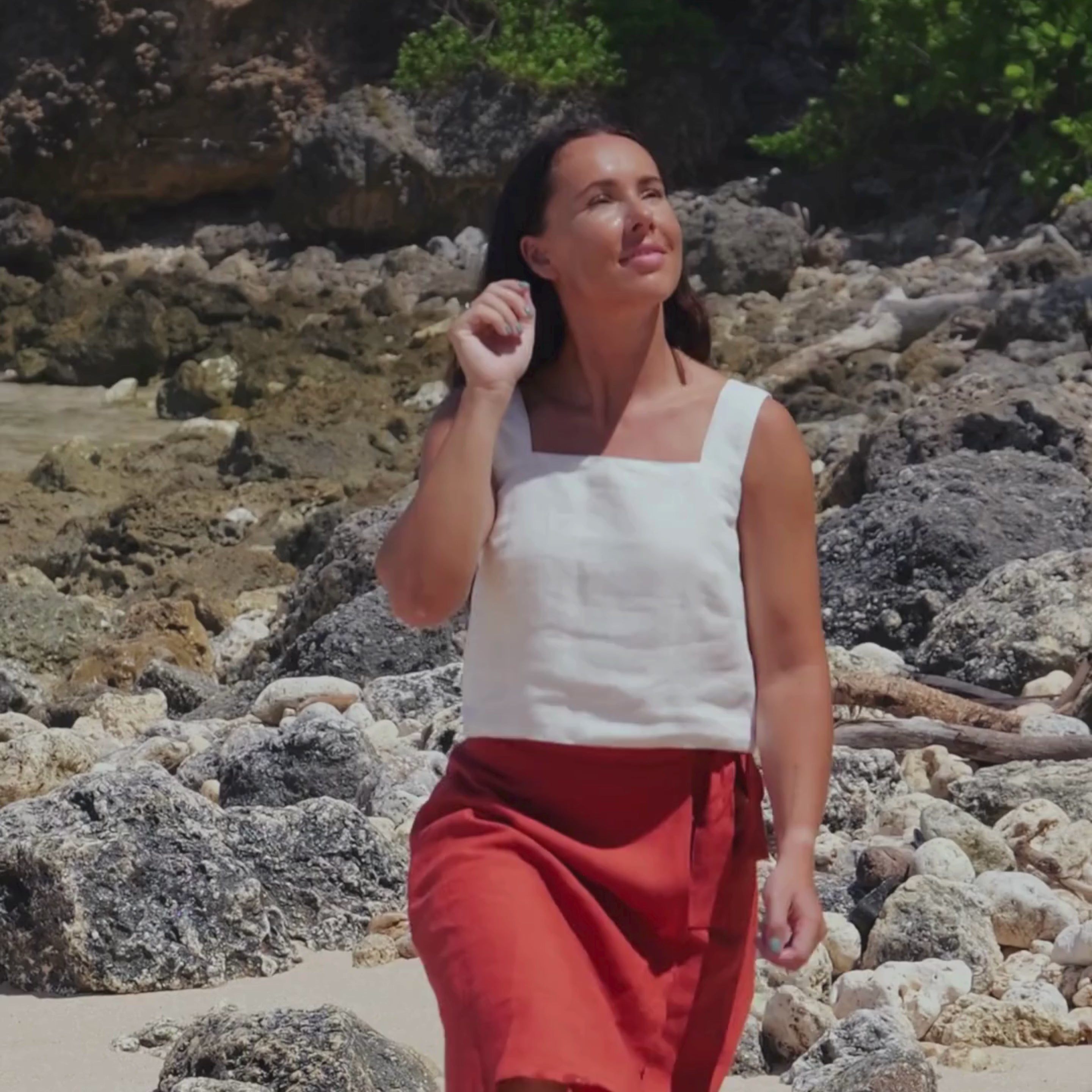 A woman with long, dark hair is featured in a short video clip. She is wearing a pure white menique linen top with wide straps. She is standing near a light-colored wooden fence, looking towards the camera with a slight smile. Green foliage is visible in the background, suggesting an outdoor setting.