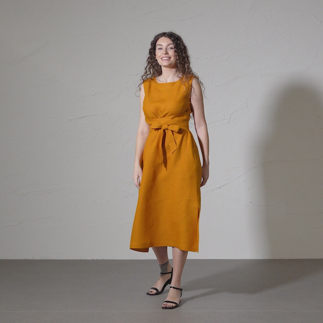 A woman with curly brown hair smiles and models a sleeveless, spicy yellow linen dress with a tie waist against a textured gray backdrop in a short video.