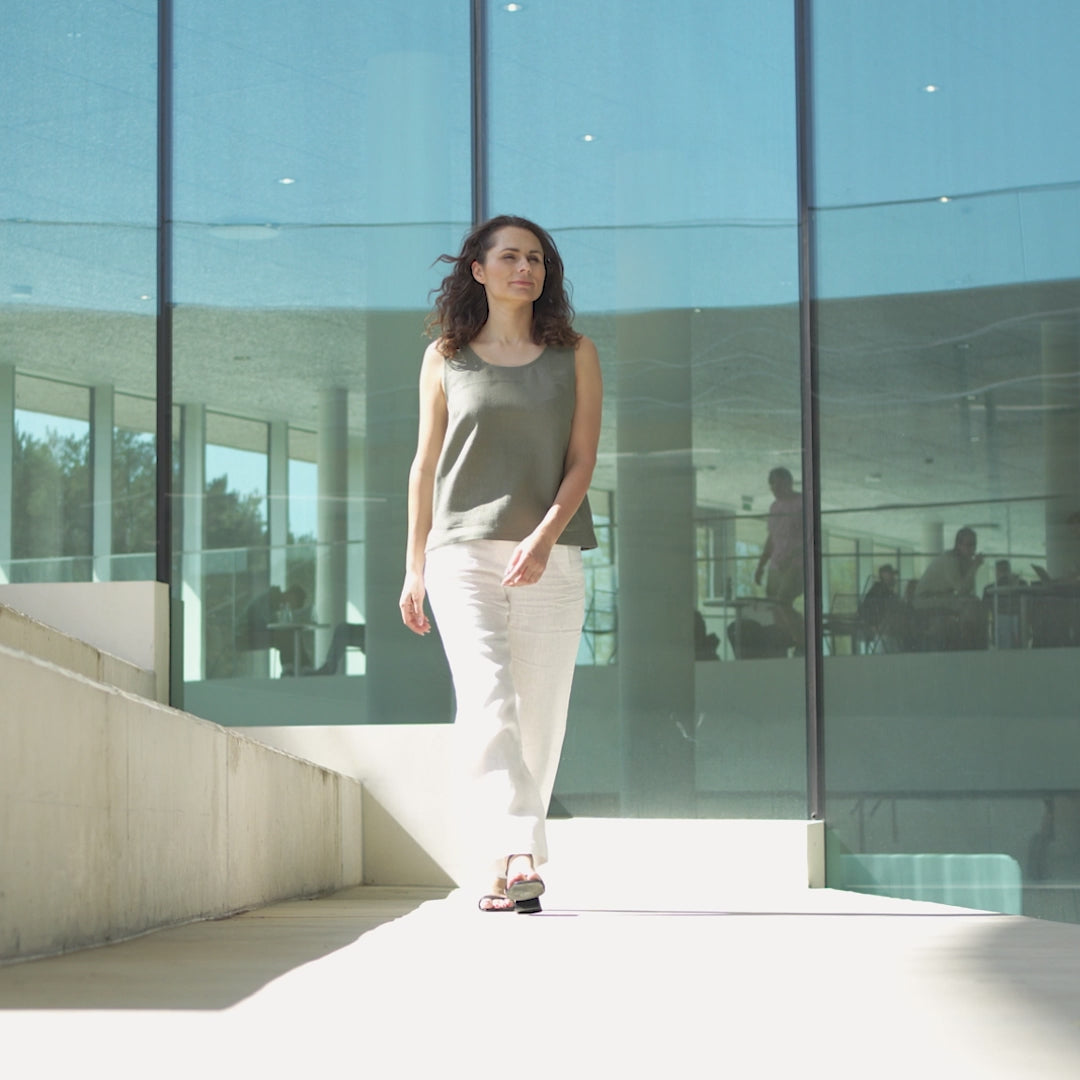 A woman with curly dark hair walks toward the camera, modeling a sleeveless stone green linen top and white pants, in a short video set outdoors against a modern glass building reflecting a bright sky.