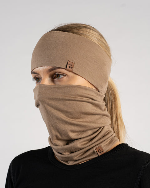 A woman looking slightly to the left, wearing a matching beige headband and a neck gaiter pulled up over her nose and mouth, covering the lower half of her face. She has light eyes and blonde hair pulled back into a ponytail. She is wearing a black top. Both the headband and the gaiter feature a small, rectangular brown leather logo tag. The background is a solid light grey.