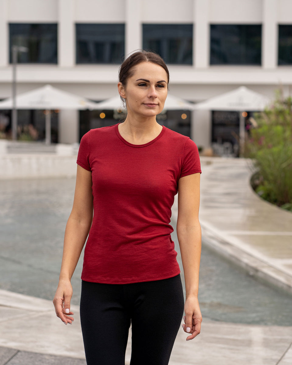 Vrouw draagt een Menique Royal Cherry t-shirt van merinowol en zwarte broek, staand in een buitenomgeving met gebouwen en parasols op de achtergrond.
