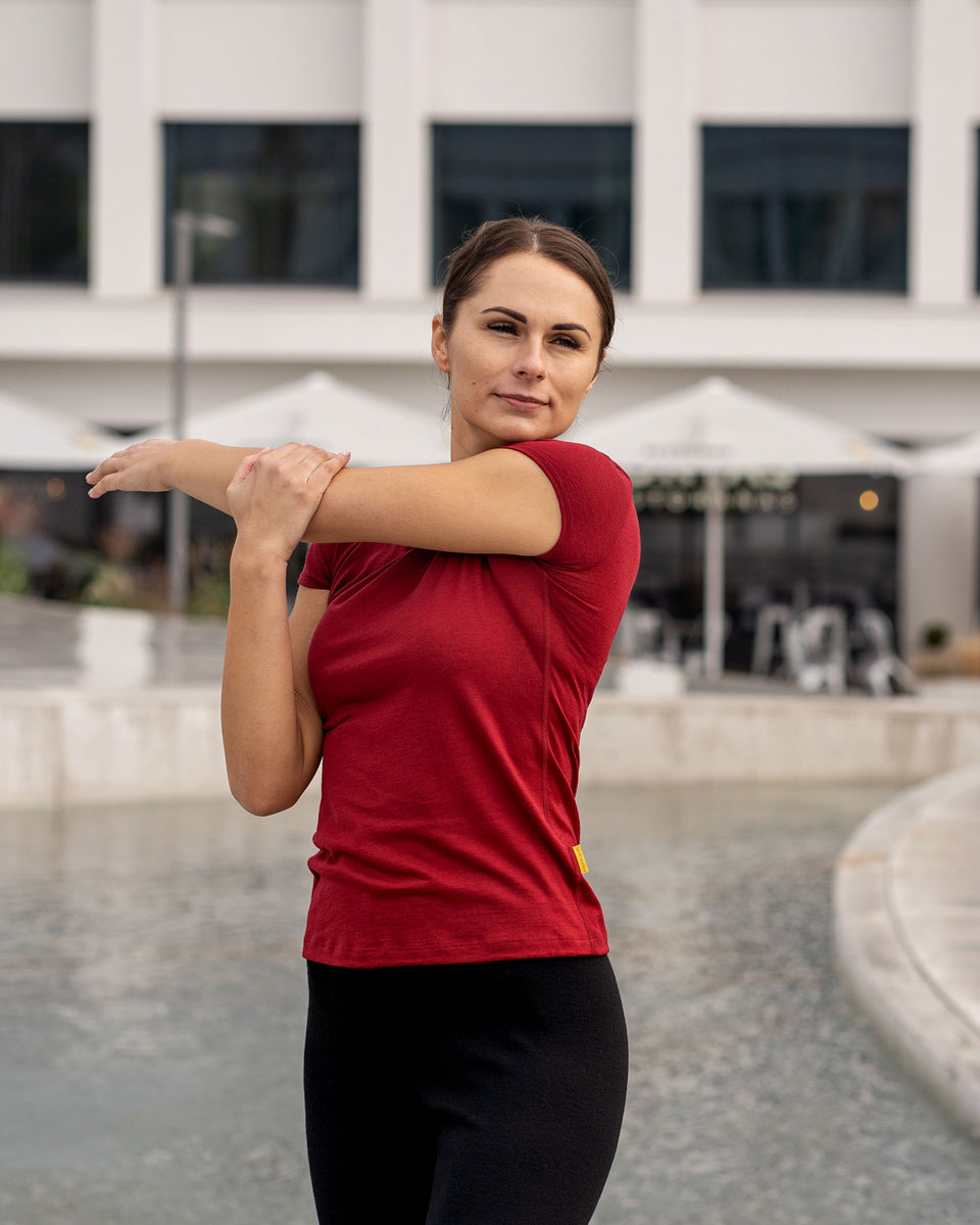 Vrouw in een merinowollen Menique T-shirt in de kleur koninklijk kers rood die buiten rekt met een gebouw op de achtergrond