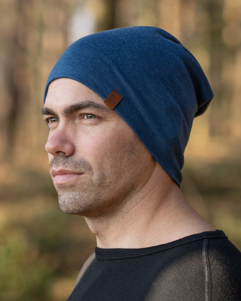 A man standing outside during fall season, in a forest, wearing the menique Men's Merino Beanie Hat in Denim Color.
