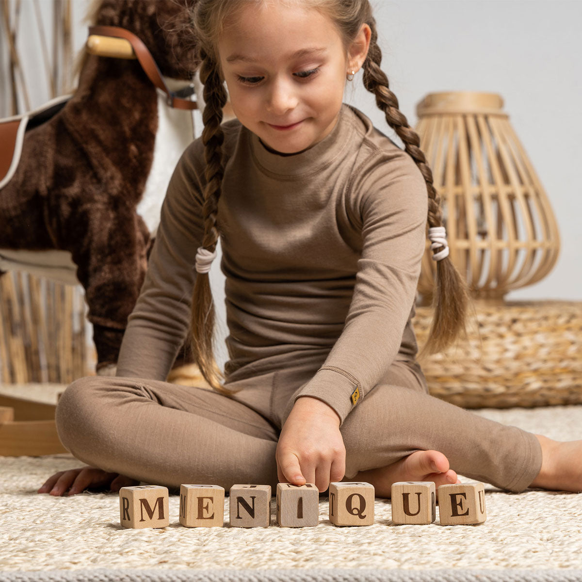 Een meisje, met een teddybeer in haar handen, draagt de menique Merino 160 Broek in beige kleur. Ze staat op een bed en lacht.